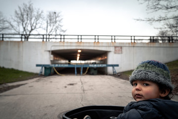 Young baby boy sits in stroller looking at camera wearing knit hat and jacket with police barricades blocking an underpass leading to lakefront in background in Chicago during the COVID-19 outbreak.