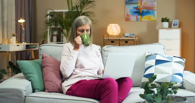 Beautiful joyful Caucasian senior woman with glasses typing on computer while sitting on sofa at home and drinking coffee. Pretty isolated old lady texting on laptop and sipping hot drink Rest concept