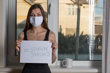 European girl wearing medical face mask holding paper with hashtag Stay At Home in her house. Patient isolated to prevent infection of pandemic, epidemic Covid-19. Stop virus