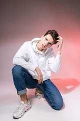 Young caucasian male model posing while sitting on the floor in the studio. Teenage boy in stylish clothes on a pink background. Jeans guy, white hood and sneakers