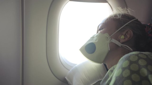 Woman travel caucasian at plane with wearing protective medical mask. Girl tourist at aircraft with respirator, feel bad, sneezes, sore throat and head, fever, sleeps. Coronavirus sars-cov-2 covid-19.