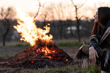 Happy young woman sitting near the evening midspring night campfire among field