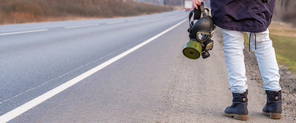 Banner. Black gas mask in the hand of a girl standing on the edge of a country highway. The girl is...