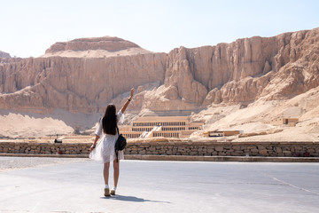 Young brunette female in white dress walks in front of part of the Mortuary temple of the Queen Hatshepsut Dayr el-Bahari or Dayr el-Bahri , Western Bank of the Nile