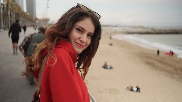 Close up shot of pretty smiling hispanic girl in red hoodie happily walking along seafront 