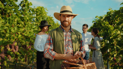 Portrait of proud caucasian farmer in strawhat smiling with joy holding large basket of grapes working during harvest in vineyard.