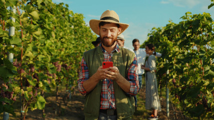 Naklejka premium Joyful bearded farmer wearing strawhat browsing fun content on smartphone during rest on grapevine alley at vineyard. Wine worker. People and technology.