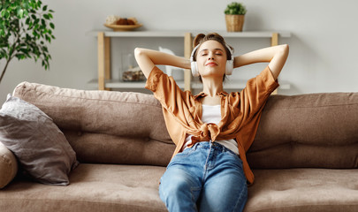 Pleased young woman in headphones listening to music while relaxing on sofa at home