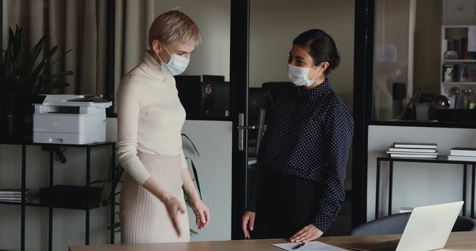 Indian and caucasian businesswomen wear medical face masks greeting with elbow bump work together in office. Two female colleagues avoid touch for coronavirus protection covid 19 spreading prevention.