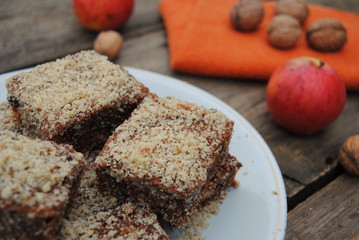 Sweet Apple Cake Slices, sprinkled with ground walnut, served in a white ceramic plate. Apples and Walnuts in the background. Close up view.