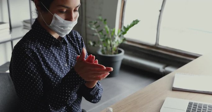 Young indian business woman wearing face mask cleaning hands with sanitizer gel at workplace. Female worker using alcohol sanitiser working from home office on computer to prevent coronavirus concept.