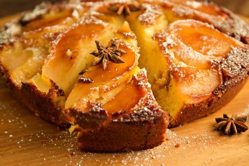 Pear upside down cake with castor sugar on a wooden board with a slice cut out.  The slice is decorated with aniseed.  A single aniseed sits on the wooden board in front of the cake.