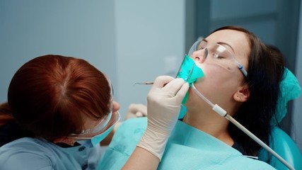 Woman in chair at stomatology clinic. Professional female dentist in protective mask and glasses treating patient's teeth using medical equipment.