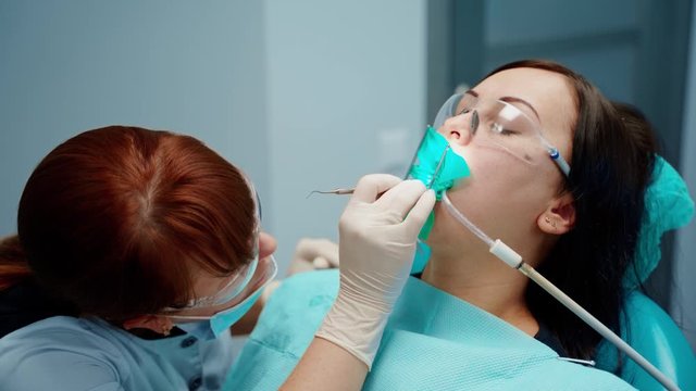 Woman In Chair At Stomatology Clinic. Professional Female Dentist In Protective Mask And Glasses Treating Patient's Teeth Using Medical Equipment.