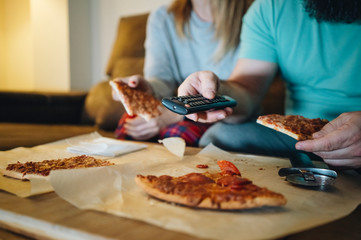 A couple of men and women eat pizza on the sofa in their living room at night while watching a movie on television. Concept of life as a couple at home.
