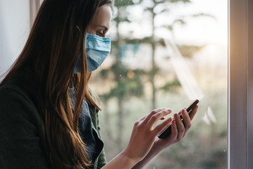 Female working distantly from home office using modern smartphone. Young woman wearing face mask sitting on window sill at home, looking at screen phone, chatting in social network. Social Distancing