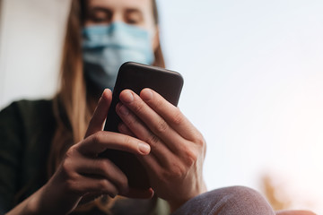 Woman in medical sterile face mask holding in hands mobile phone, close up selective focus on arms and electronic gadget, sitting on window sill at home. Concept social distancing, Home quarantine