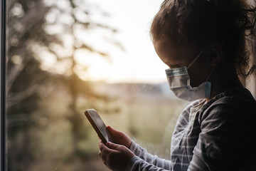 Tired little girl in medical sterile face mask sitting on window sill using modern smartphone gadget playing or browsing, upset pensive kid feeling lonely. Self isolation, home quarantine
