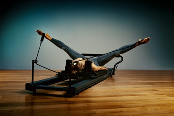 Young girl doing pilates exercises with a reformer bed. Beautiful slim fitness trainer on reformer gray background, low key, art light. Fitness concept