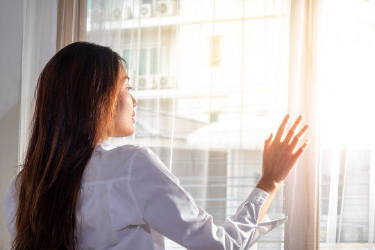 Rear View Of Relaxing Young Beautiful Asian Woman Girl Wake Up In Home Bedroom And Using Her Hand Opening Balcony Window Curtains With Sun Shining Through The Curtain To The Room In Fresh Morning.