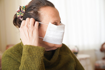 Portrait of old woman wearing surgical mask for protection against corona virus. Grandmother sitting in her living room and looking up 