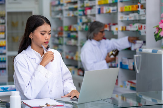 Young Asian female Pharmacist using laptop computer checking medicines and health care product inventory stock at hospital pharmacy or drugstore. Medical, pharmaceutics and personal healthcare concept