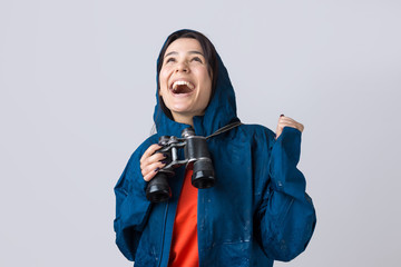 A tourist girl in a blue raincoat holds binoculars in her hands and looks into the distance, spies.