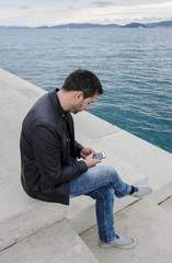 Handsome man sitting on the pier, using smartphone