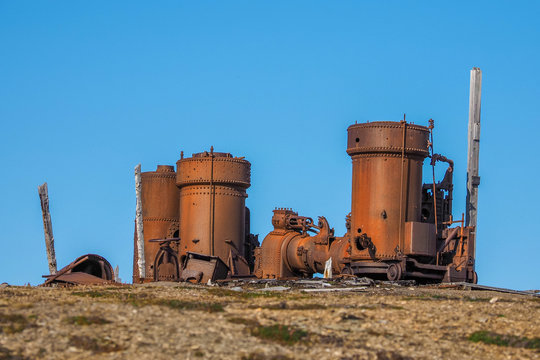 Abandoned rusty mining machinery and industrial equipment on a barren tundra hill under a clear blue sky in the historic mining area of Svalbard. Camp Mansfield Spitsbergen Ny London,