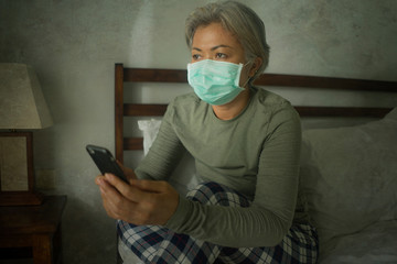 worried middle aged woman 50s with grey hair and protective mask checking online news with mobile phone during covid-19 virus home lockdown quarantine