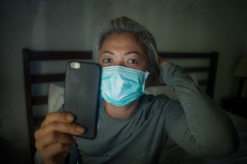 worried middle aged woman 50s with grey hair and protective mask checking online news with mobile phone during covid-19 virus home lockdown quarantine