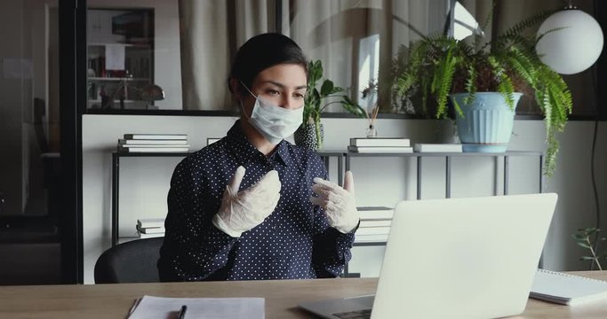 Young indian business woman wearing medical gloves and face mask communicating by video call on laptop computer. Online conference distance remote home office during coronavirus covid 19 quarantine.