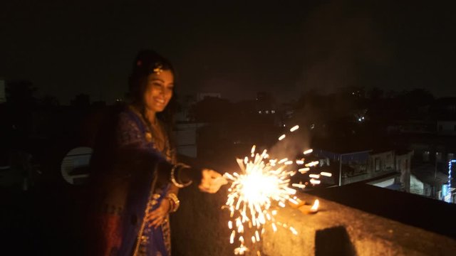 Young and beautiful Indian Bengali woman in Indian traditional dress is celebrating Diwali with fire crackers on a rooftop in darkness. Indian lifestyle and Diwali celebration
