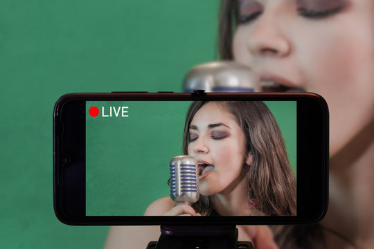 Focus on live view on smartphone on tripod, girl singing with microphone image on back screen with blurred scene in background. Social media live streaming concept