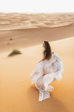 Portrait Of Bride Woman In Amazing Wedding Dress In Sahara Desert, Morocco. Warm Evening Light, Beautiful Pastel Tone, Sand Dunes On Horizon. View From Behind.