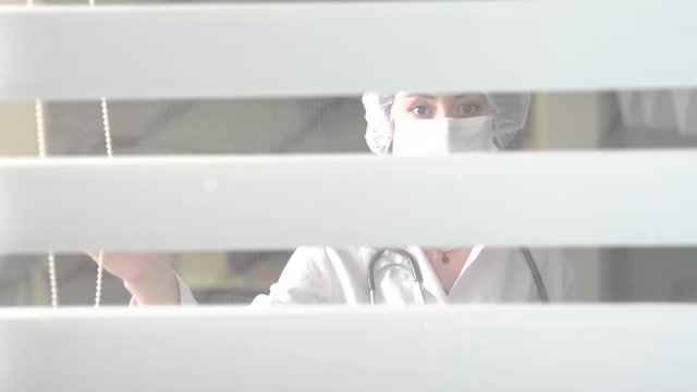 A Female Doctor With Mask Is Looking Outside Curiously From Window At The Office.