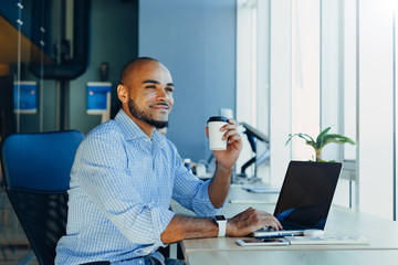 Confident young african american businessman working on laptop and talking on a cell phone while sitting at his workplace in startup office