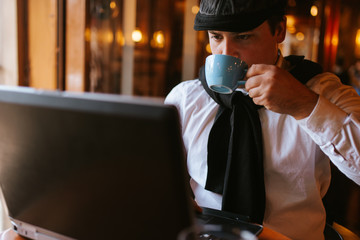 A young handsome caucasian businessman with a cap sits at a cafe drinking coffee in front of a laptop