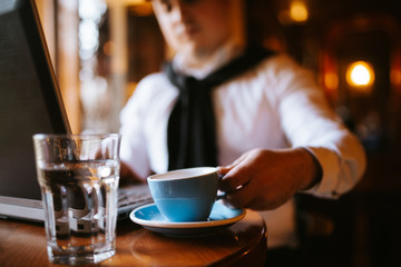 A young handsome caucasian businessman with a cap sits at a cafe drinking coffee in front of a laptop