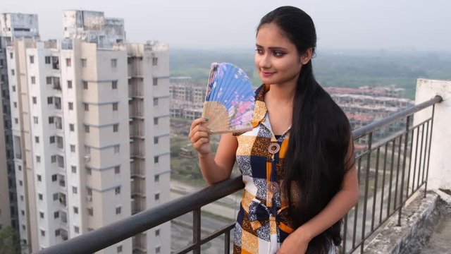 An young and attractive Indian Bengali brunette girl in western dress enjoying with a Chinese fan on a balcony/rooftop in urban background. Indian fashion portrait and lifestyle.