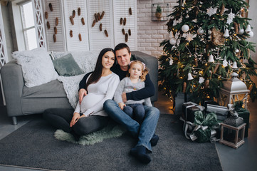 A man, a pregnant woman and a daughter are sitting and hugging on a woolen rug in a beautiful interior, in a modern studio with New Year's decorations. A happy family. Photography, concept.