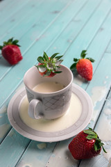 Red ripe strawberries and a white coffee Cup, on a turquoise wooden table. The berry falls into the milk. Frozen movement. Splashes from the Cup.