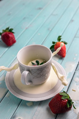 Red ripe strawberries and a white coffee Cup, on a turquoise wooden table. The berry falls into the milk. Frozen movement. Splashes from the Cup.