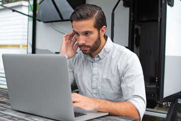 A man wearing a long sleeve grey button down business shirt with brown slicked comb over hair working on laptop on a picnic table remotely near his camper able to avoid the busy society work community