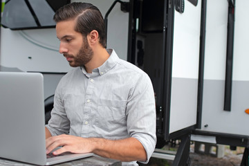 A man wearing a long sleeve grey button down business shirt with brown slicked comb over hair working on laptop on a picnic table remotely near his camper able to avoid the busy society work community