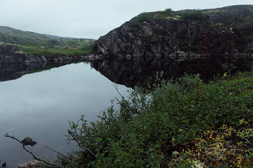 Severe north grey arctic landscape slope of granite rocks and quiet dark lake with lush green meadow in haze in overcast weather.