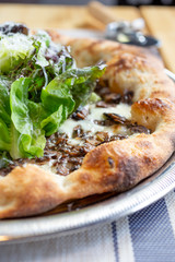 A closeup view of a rustic pizza pie in a restaurant or kitchen setting, featuring toppings as mushrooms and gem lettuce greens. 