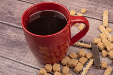 A red ceramic tea mug, cookie sticks with chocolate and white icing, and pieces of brown cane sugar on a wooden background. Close up.