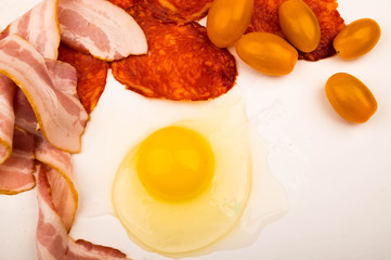 A broken chicken egg without a shell, slices of bacon and sausage and tomatoes on a white background. Close up.