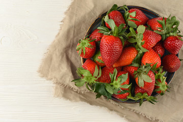 Fresh ripe strawberries in ceramic plate on white wooden table. Linen towel under the plate. Top view, copy space. 